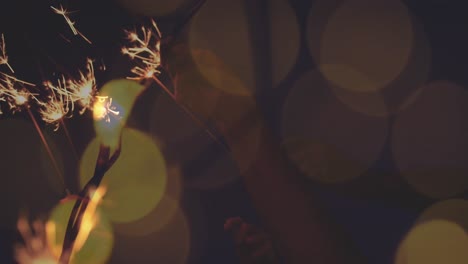 animation of hands holding new year sparklers over orange and yellow light spots against night sky