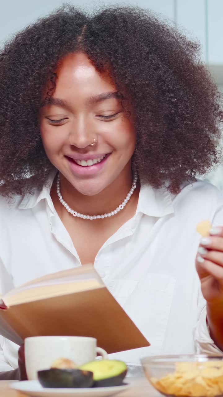 Young african american woman laughs reading book and eats corn sits in kitchen