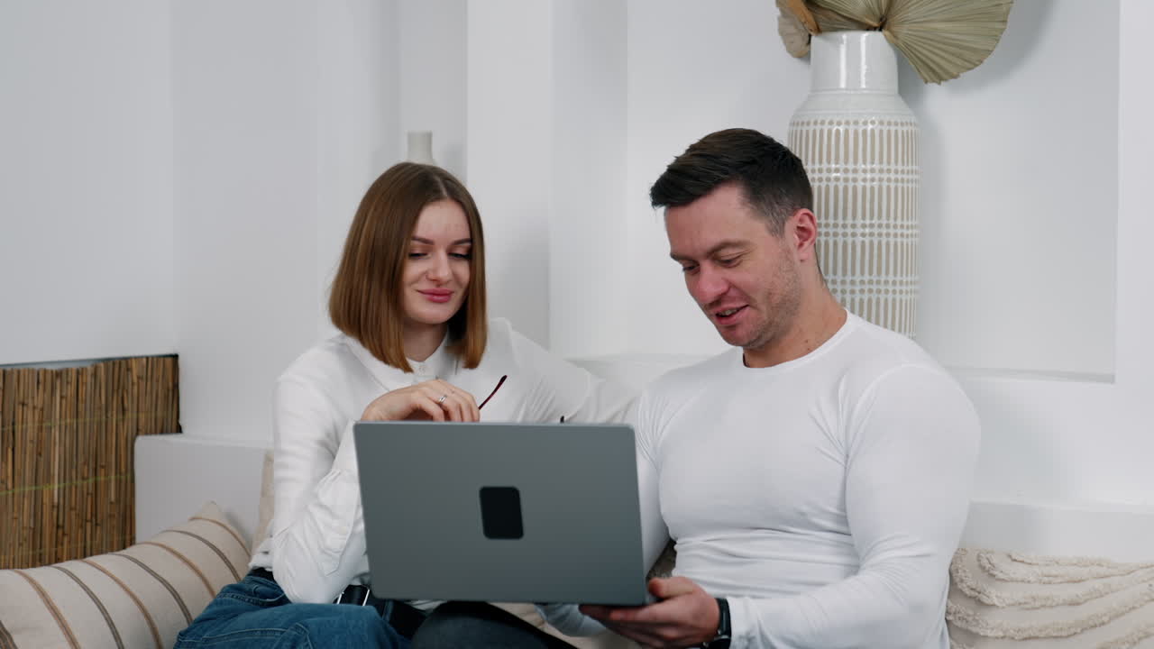 Caucasian couple sitting at sofa talk looking at laptop screen. Remote employment in comfortable atmosphere.
