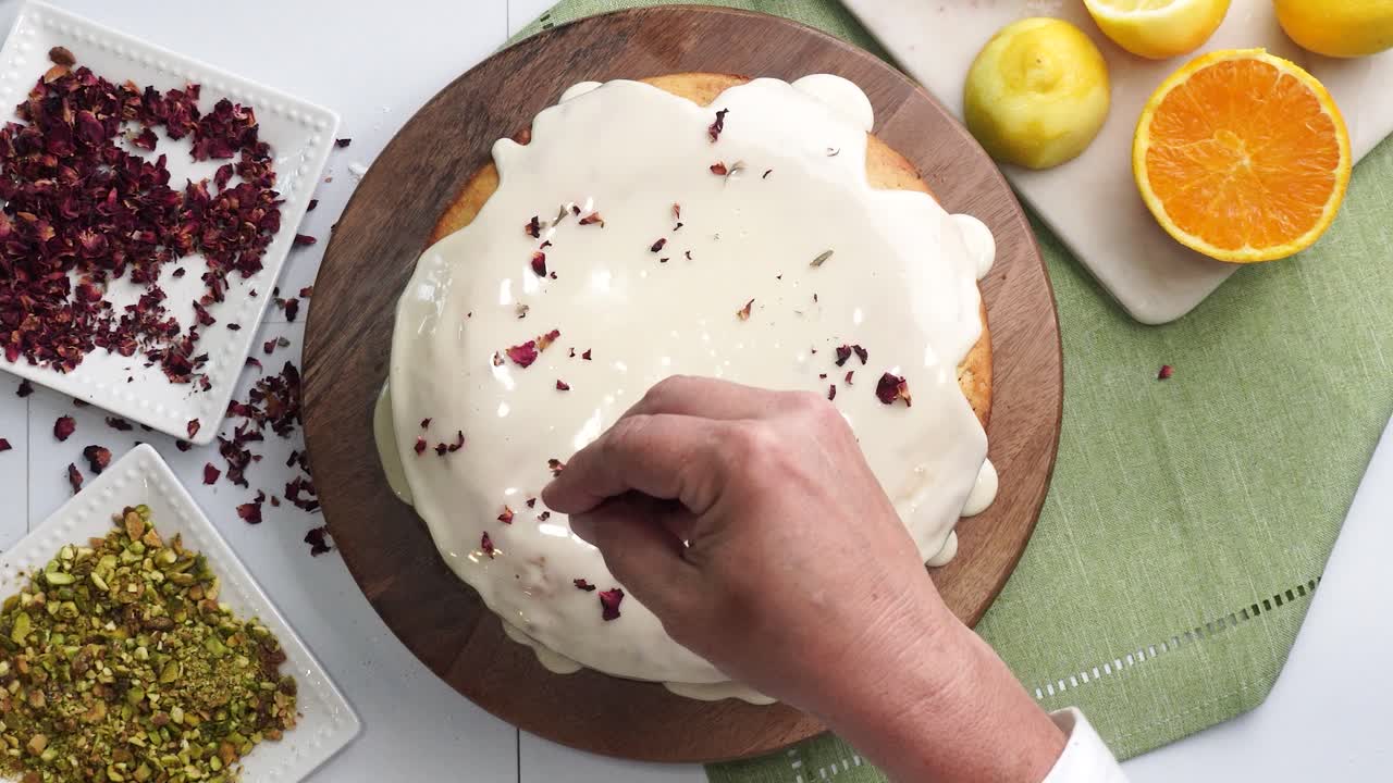 Baking a dessert called Persian Love Cake made with rose petals and pistachios. Cake with white glaze frosting. Decorating the top with red rose petals. Beautiful cake for tea time or celebration