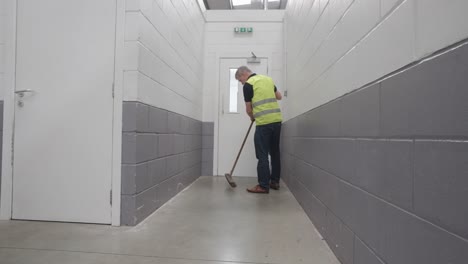 male wearing yellow high vis vest brushes floor of building, locked off