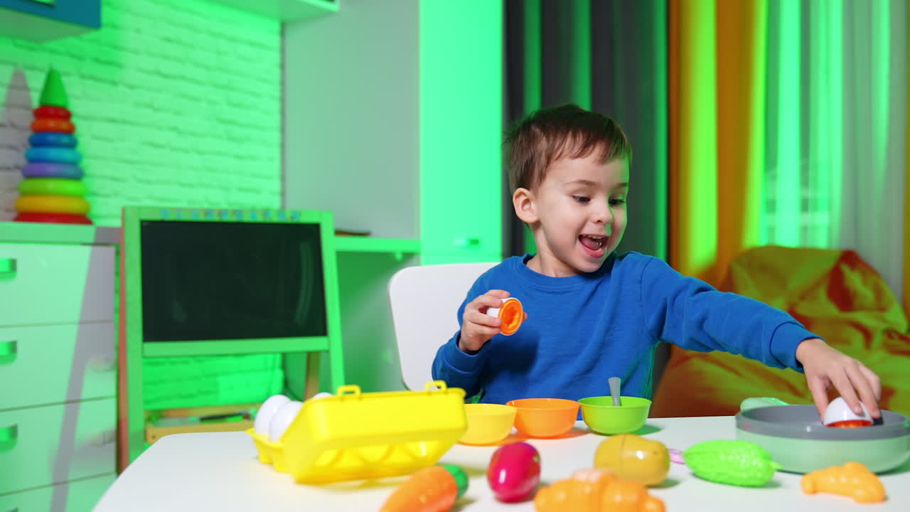 Adorable toddler boy sits at the desk indoors. Happy child plays with plastic crockery and vegetables. Educating game at home.