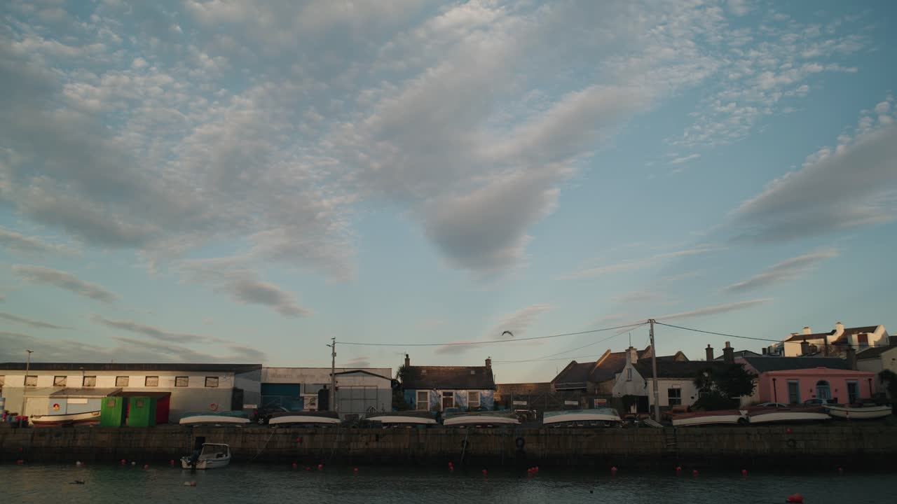 A locked-off shot at Bullock Harbour in Dublin, Ireland. A cyclist rides past waterfront houses as boats are moored along the harbor under a soft blue sky at sunset.