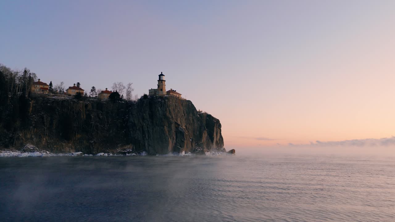 Una stabile ripresa da drone del faro di Split Rock su un'alta scogliera durante un'alba invernale colorata con nebbia di mare che sale dal Lago Superiore