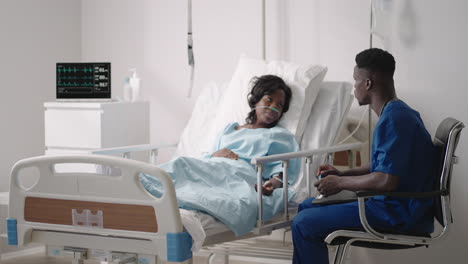 A-black-male-doctor-is-talking-to-a-black-female-patient-lying-on-a-hospital-bed-and-connected-to-an-acid-supply.-The-attending-physician-discusses-with-the-patient-in-the-hospital-the-treatment