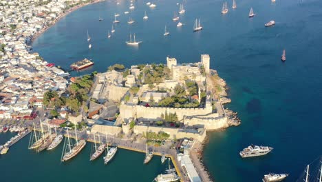 Luftaufnahme-Der-Burg-Von-Bodrum,-Umgeben-Von-Der-Blauen-Ägäis-Mit-Vielen-Im-Wasser-Verankerten-Booten-An-Einem-Sonnigen-Sommernachmittag-In-Mugla,-Türkei,-Während-Des-Sonnenuntergangs