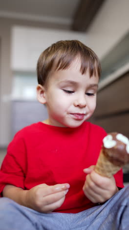 Cute toddler boy sits on the floor eating ice-cream. Lovely baby pinches off the pieces of waffle cone. Vertical video.