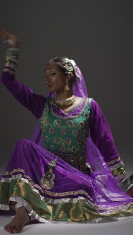 vertical video of female kathak dancer performing dance wearing traditional indian dress seated on floor in spotlight 2