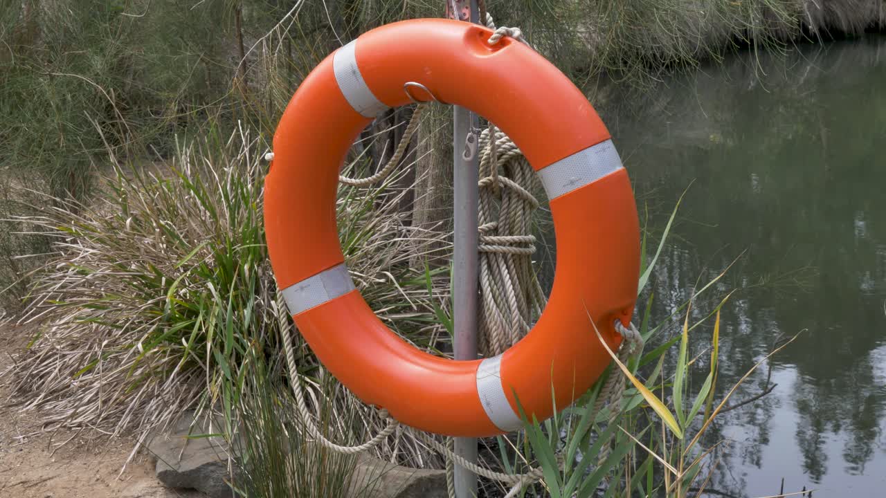 Slow motion view of orange lifebuoy ring safety flotation device with rope on pole beside river lake inlet embankment and bushland forest plants at national park in South Coast NSW Australia rescue