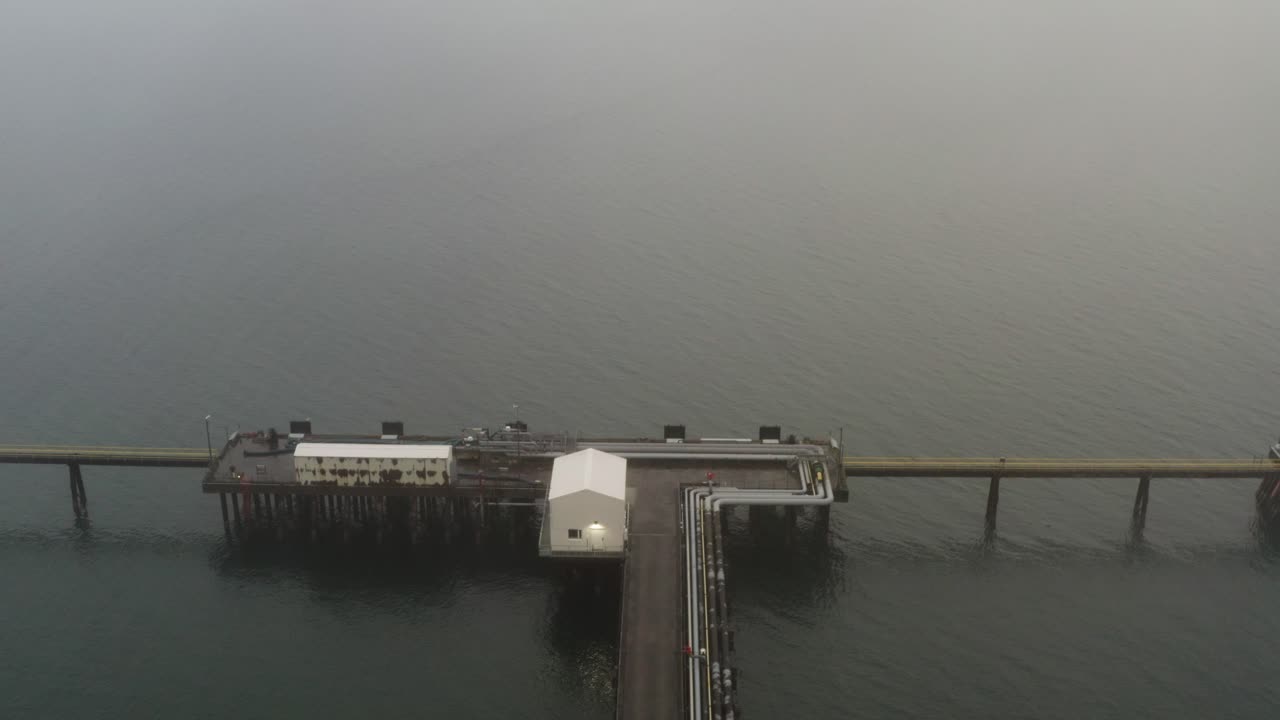Long wooden pier extending over still water in foggy Hvalfjörður Iceland with small white buildings and distant hills fading into mist under overcast daylight