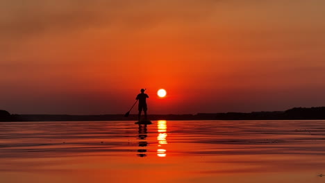 summer evening time on the calm river. lonely male silhouette on a sup board floating on water. orange sunset background.