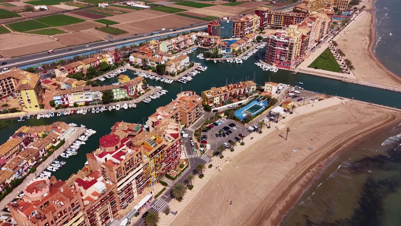 Port Saplaya marina i Alboraya, Valencia, Spanien, der viser farverige bygninger ved vandet, fortøjede både, sandstrand og omkringliggende landbrugsjord set fra oven.