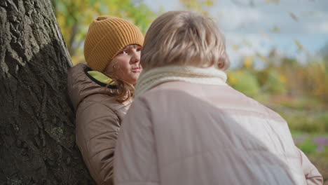 woman approaches daughter sitting on tree trunk and lovingly hands over autumn leaf before leaning in to give warm peck in calm park filled with vibrant fall foliage and soft cloudy sky