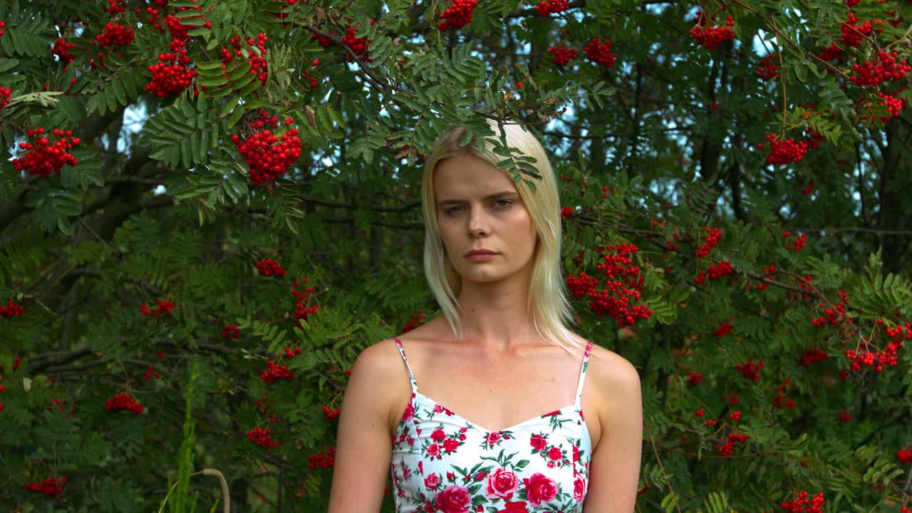 붉은 열매가 달린 나무 아래 중립적인 표정으로 서 있는 여자
