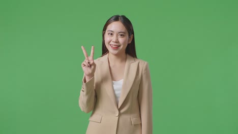 an asian business woman showing gesture peace and smiling while standing on green screen in the studio