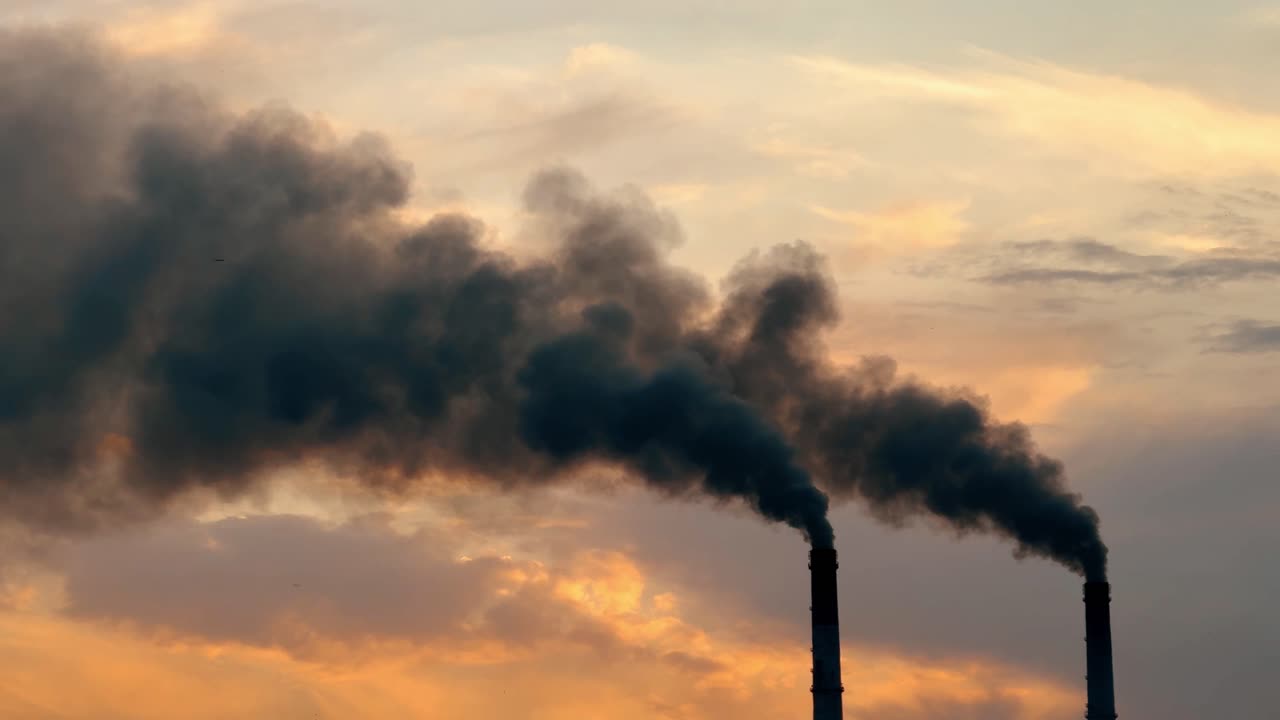 Dei fumaioli emettono fumo al tramonto in una zona industriale. Due fumaioli producono un denso fumo stagliandosi contro un cielo colorato al tramonto, vicino a una fabbrica.