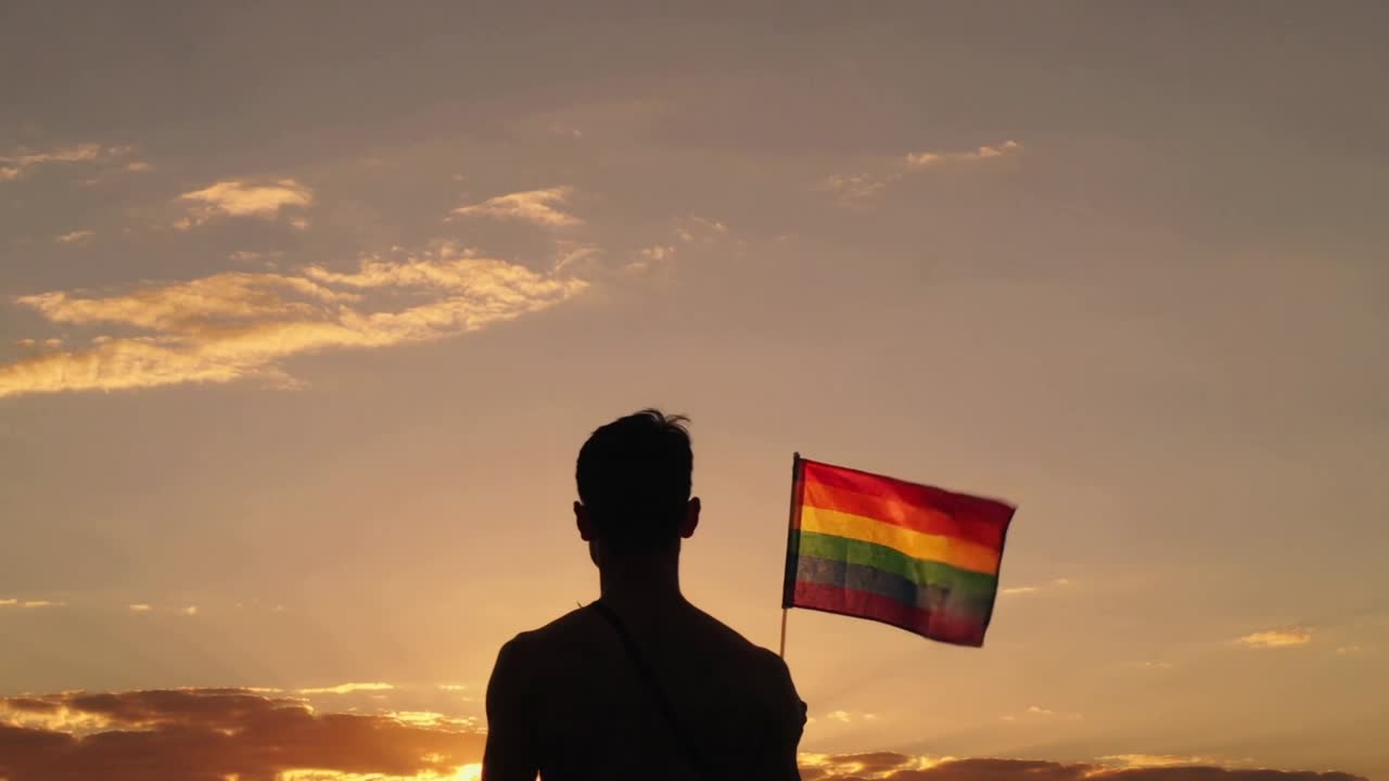 Il vento gonfia una bandiera arcobaleno del pride, un uomo a torso nudo ne aggiusta la presa, mentre il tramonto evidenzia l'orgoglio e un cinturino.