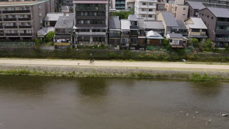 Vista-Aérea-Sobre-Ciclismo-Femenino-En-Bicicleta-A-Lo-Largo-Del-Río-En-El-Distrito-De-Edificios-De-La-Ciudad-De-Kyoto