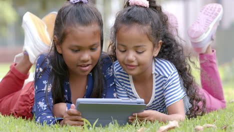 lying on grass, two girls using tablet together, enjoying outdoor activity