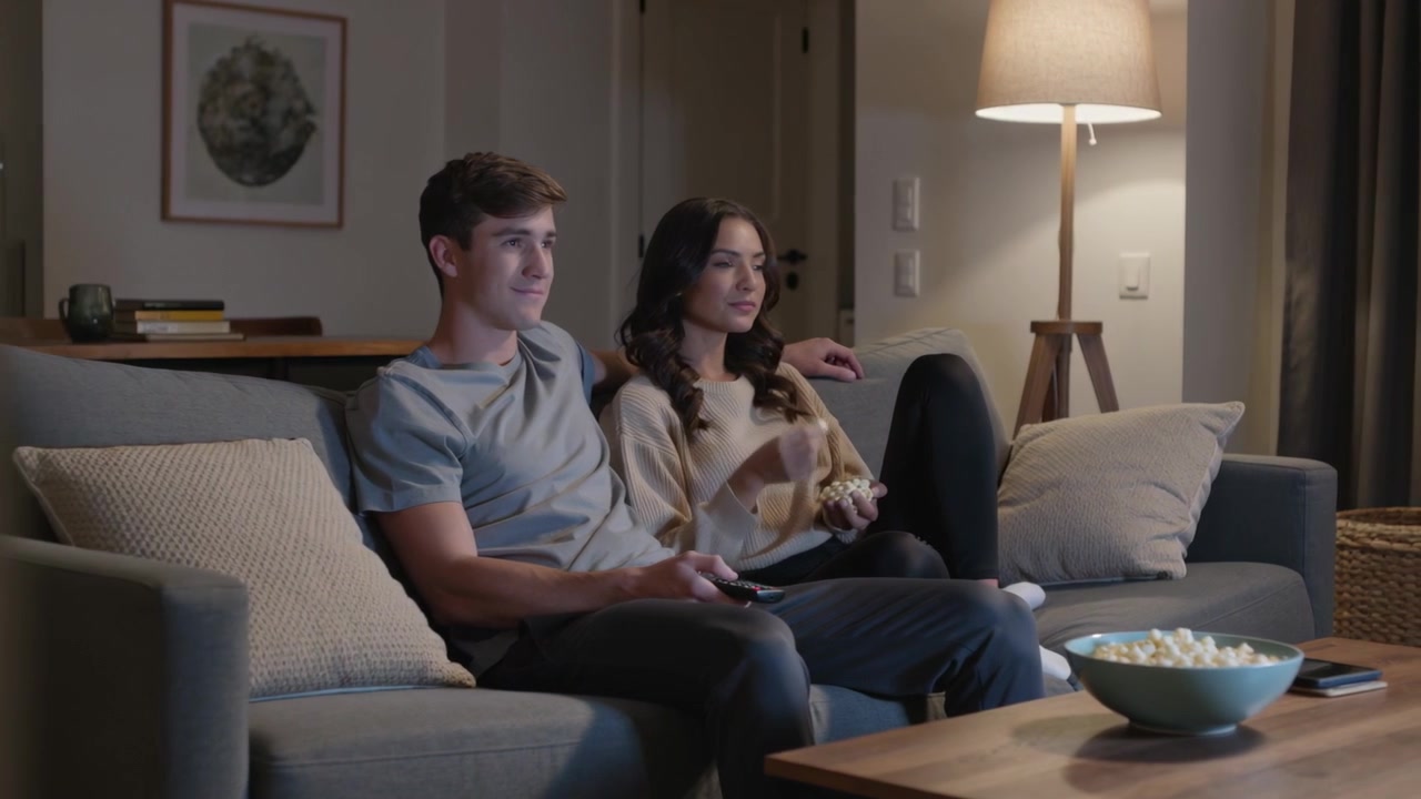 La telecamera si allontana rivelando una coppia rilassata che guarda un film a casa per rilassarsi, con popcorn e telecomando.