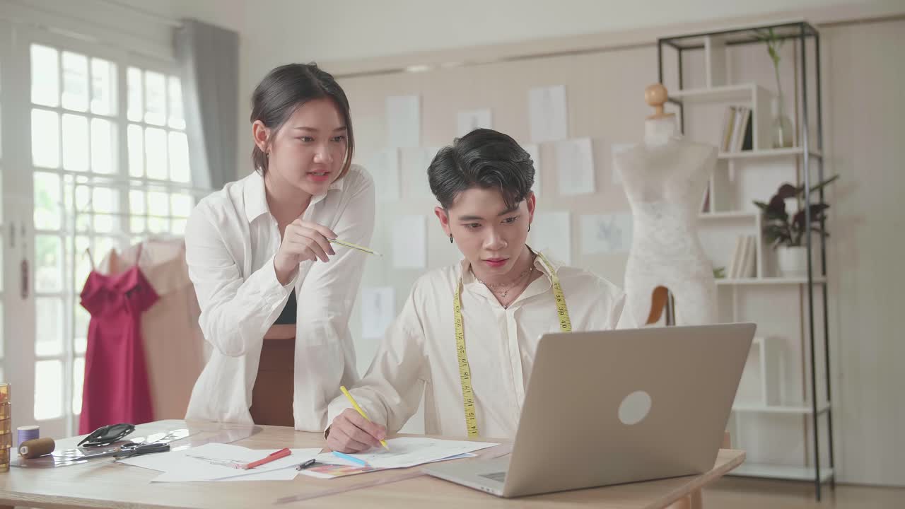 นักออกแบบแฟชั่นสองคนกำลังทำงานกับแล็ปท็อปและร่างภาพที่สตูดิโอออกแบบแฟชั่น