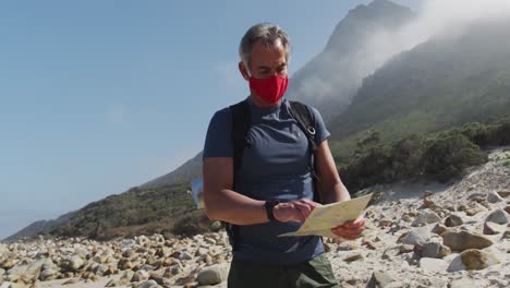 Älterer-Wanderer-Mit-Gesichtsmaske-Und-Rucksack,-Der-Beim-Wandern-Am-Strand-Karte-Liest.