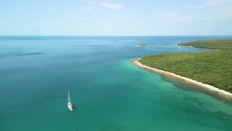 Weißes-Segelboot-Im-Ruhigen-Mittelmeer-Vor-Einer-Paradiesischen-Insel-Mit-Türkisblauem-Meer-Und-üppiger-Grüner-Vegetation