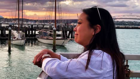 a smiling pretty asian woman gazes peacefully at a sailboat harbor and water at sunset