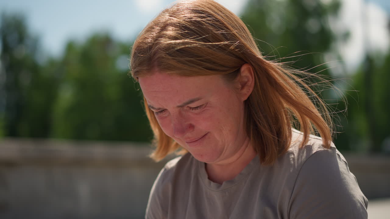 Emotional woman with windblown hair stands under bright clear sky, eyes lowered in thought, gentle sunlight highlighting face, conveying deep reflection