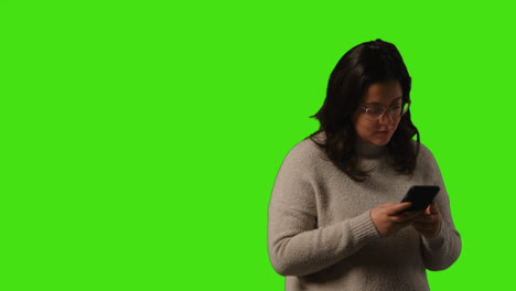 studio shot of young woman using mobile phone to text and scroll through internet standing against green screen background 1