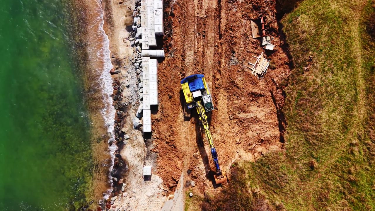 Gravemaskine bygger kystdige ved Sortehavskysten. Droneoptagelse ovenfra af betonblokke, rød jord og turkis vand langs stranden
