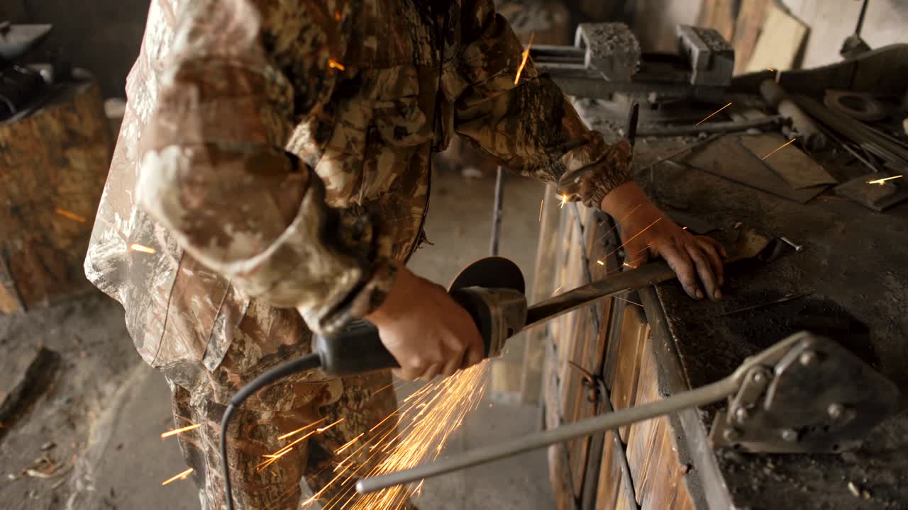 joven barbudo usando un molinillo de ángulo para trabajar en una pieza de metal en el taller de herrería, moliendo chispas volando alrededor