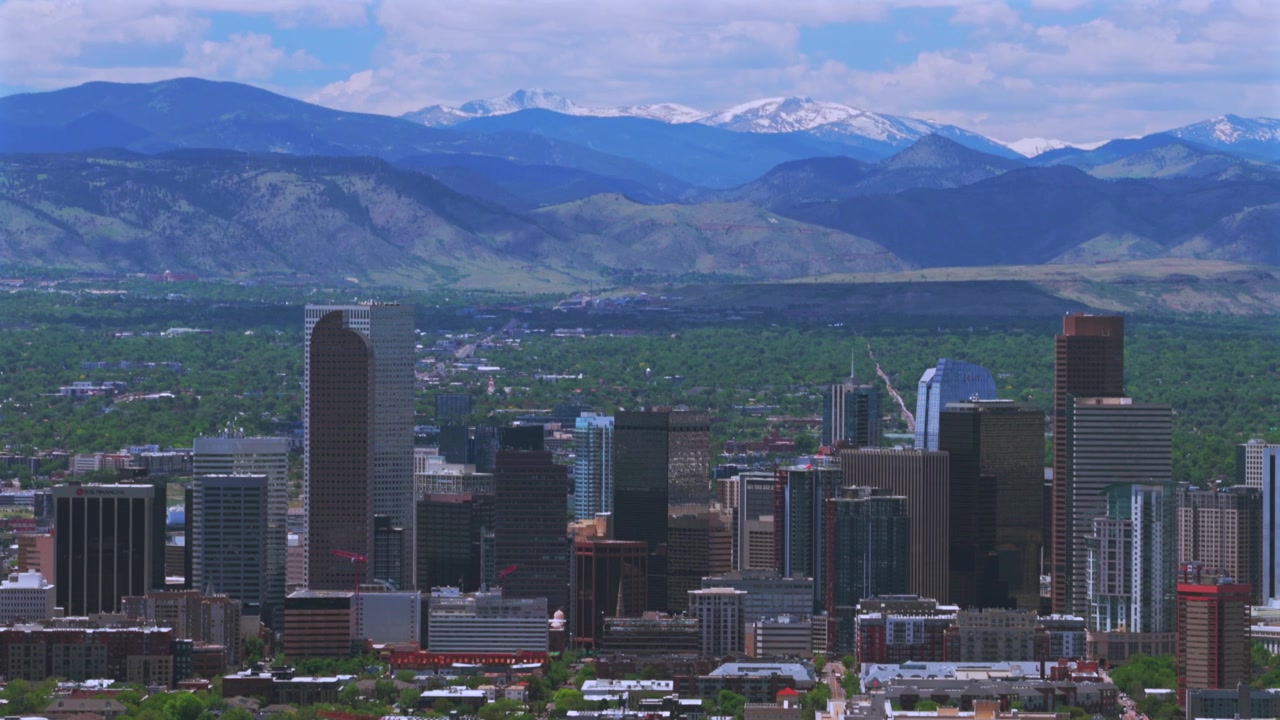 Veduta aerea con drone del centro di Denver, Colorado, con lo skyline della città, grattacieli e il paesaggio delle Montagne Rocciose della Front Range coperte di neve. Primavera, Indian Peaks, cielo blu mattutino con nuvole, il drone si muove in cerchio verso sinistra.