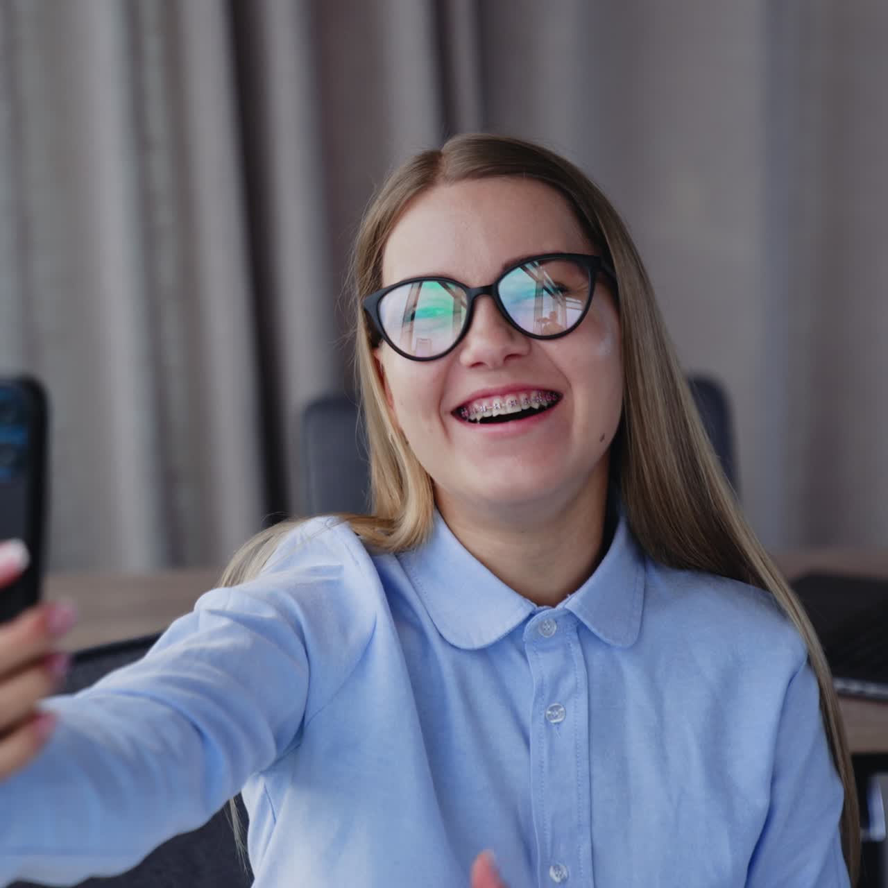 Young office employee taking selfie on her phone. Smiling lady making signs with her hand and taking pictures. Blurred backdrop