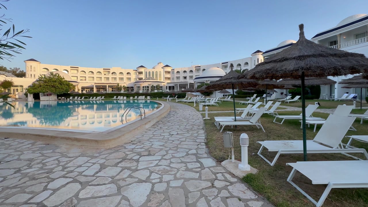 Gün doğumunda, beyaz şezlonglar ve hasır şemsiyeler, beyaz Akdeniz tarzı binaların yanındaki boş bir tatil köyü havuzunun kenarında sıralanmış. Seyahat veya konaklama temaları için kullanışlıdır. Tunus.