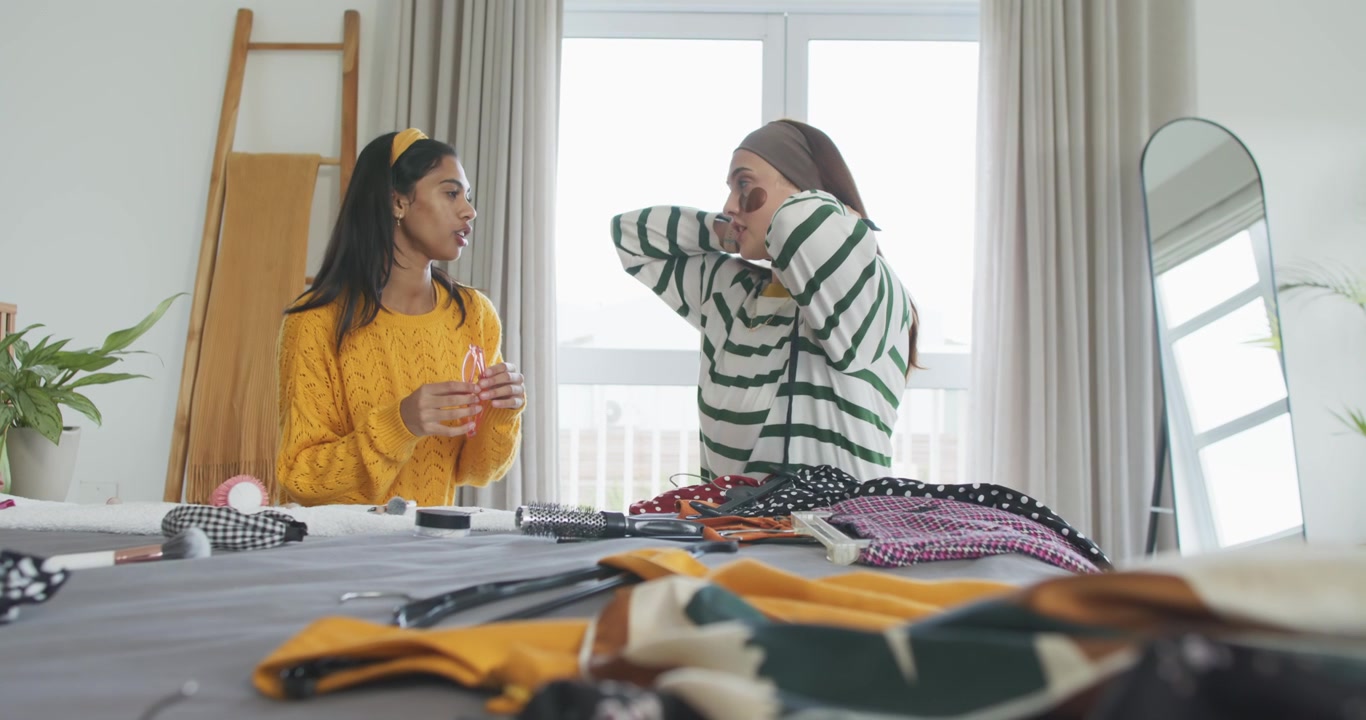 Amiche di diverse etnie mostrano un barattolo e sistemano i vestiti sul letto, provando patch per la cura della pelle e chiacchierando.