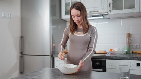 joven con un elegante vestido beige golpeando ingredientes en un cuenco blanco mientras se prepara para hornear, cocina moderna con refrigerador de acero inoxidable, armarios blancos y artículos de horneado en el fondo