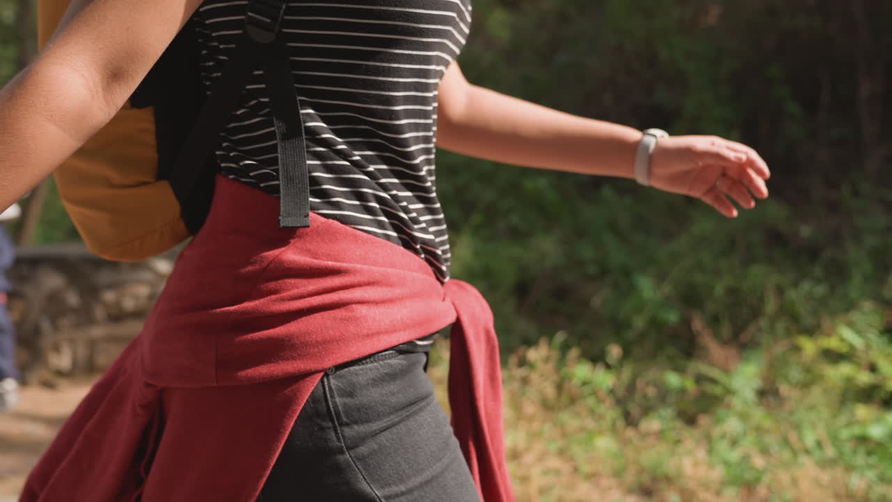 Donna di colore che cammina con maglione rosso annodato e spallina di zaino gialla, primo piano di vita e mano, stile urbano casual, famiglia che passa sullo sfondo, bracciale d'argento che cattura la luce del sole, scatto spontaneo