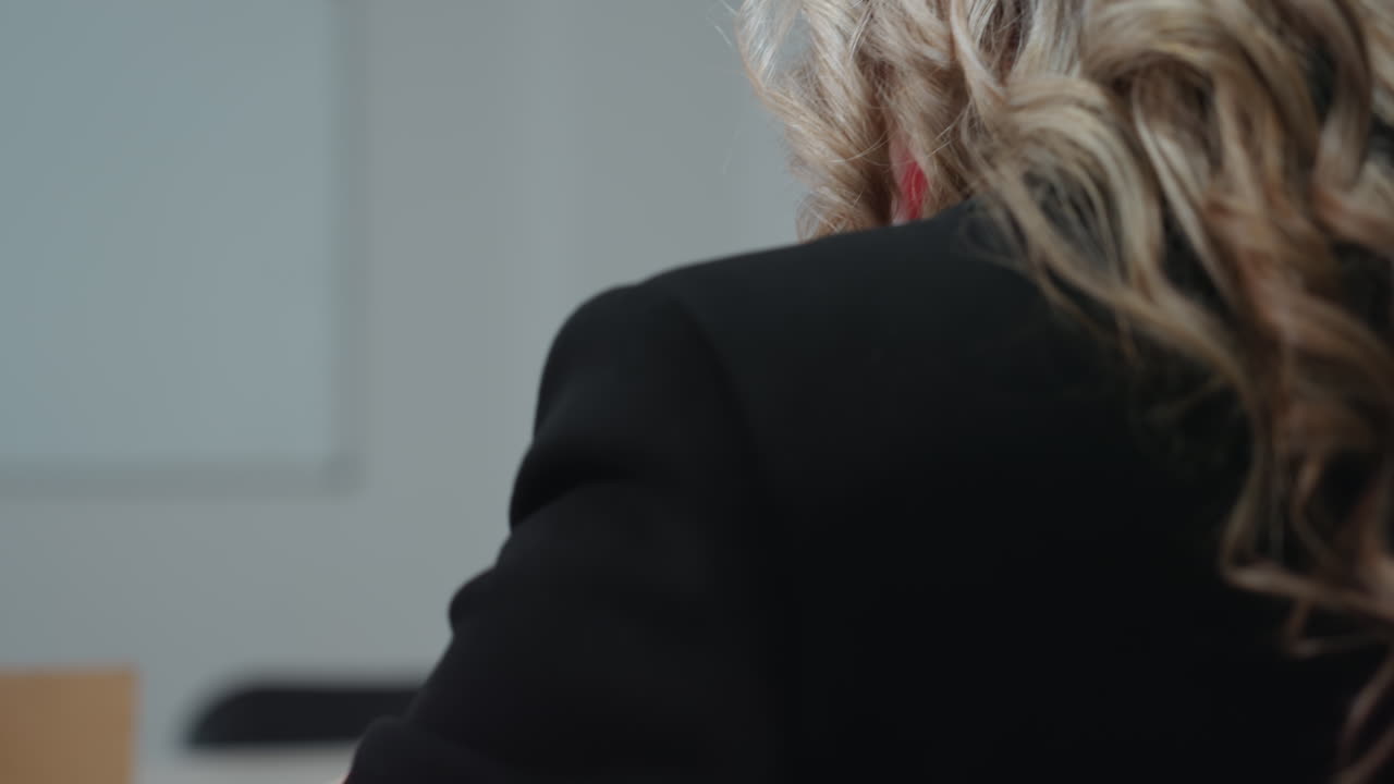 Rear view of businesswoman in black blazer opening laptop at desk with document file and smartphone nearby, ready to begin work in modern office setting