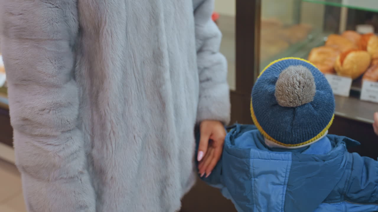 Kaukasisk mamma och son vid bageridisken, mamman i grå fuskpälsjacka och stickad mössa vägleder småbarnet i blå jacka och toppmössa när de pekar på bakverk bakom glasmontern, varmt takljus.