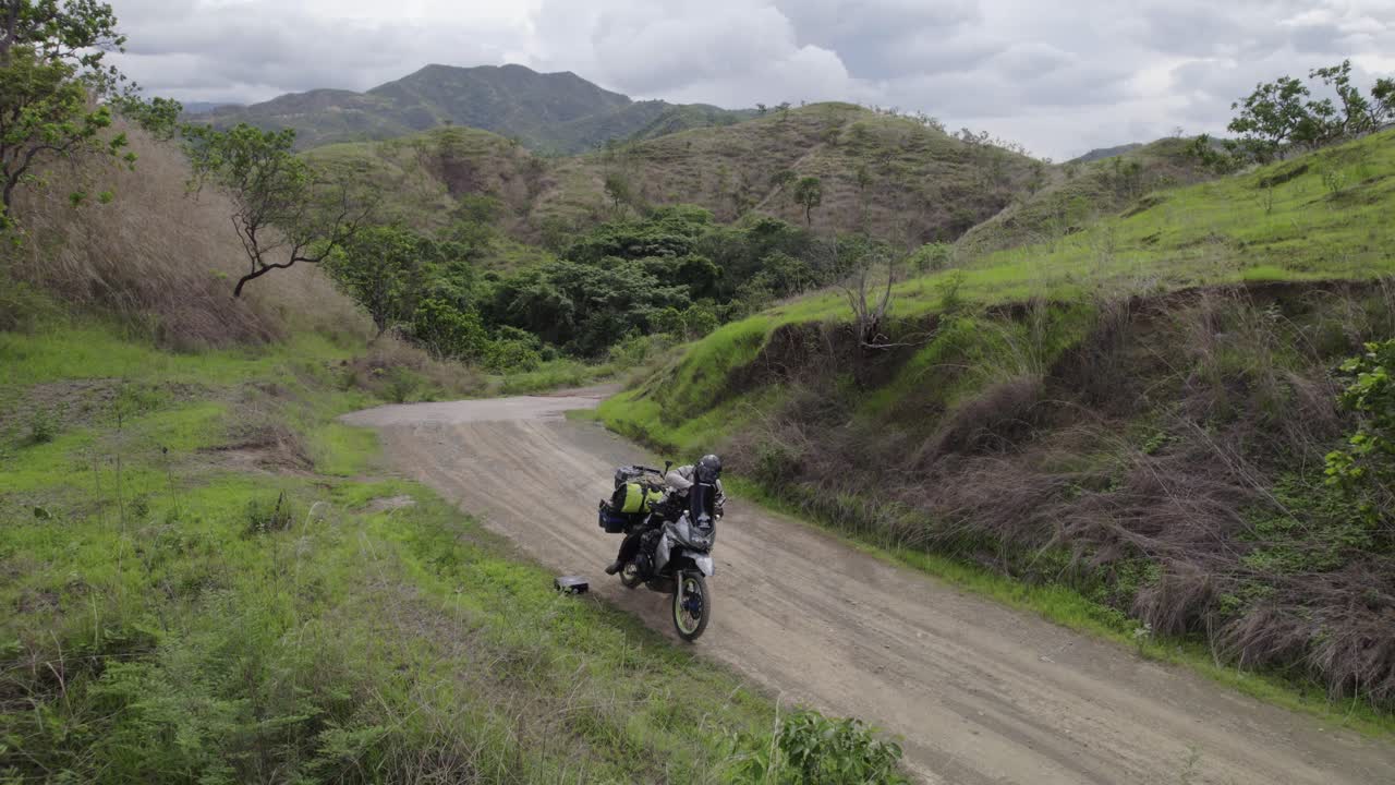Man biker riding motorcycle, adjusting jacket, starting road trip on off-road countryside of Venezuela, South America. Aerial view captures scenic landscapes, open fields, and rural adventure