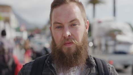 Retrato-De-Un-Joven-Barbudo-Mirando-Serio-A-La-Cámara-En-Un-Soleado-Paseo-Urbano-Frente-A-La-Playa,-Un-Hombre-Hipster-Confiado-De-Vacaciones