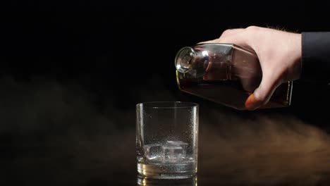 Barman pour whiskey cognac brandy from bottle into drinking glass with ice cubes on dark background