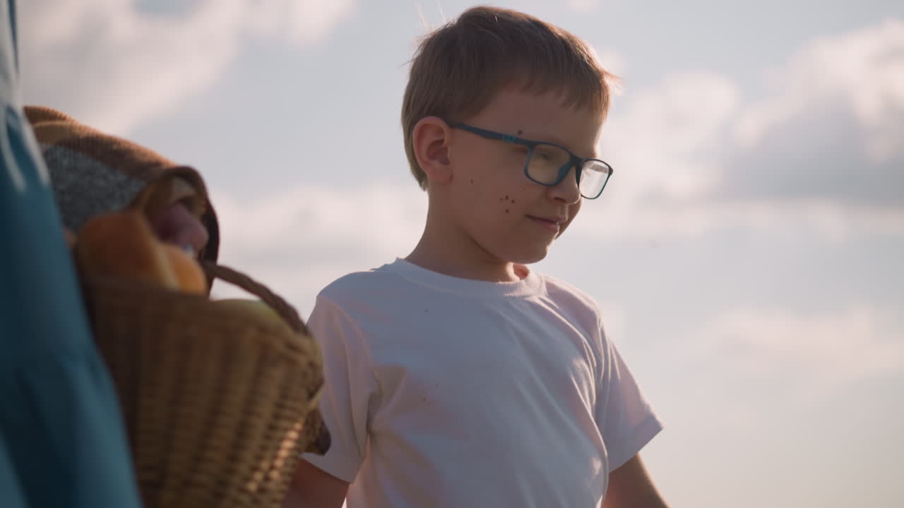 A close-up of a young boy wearing glasses and a white shirt, holding his mother's hand. The mother, partially visible, is carrying a basket filled with goods, walking