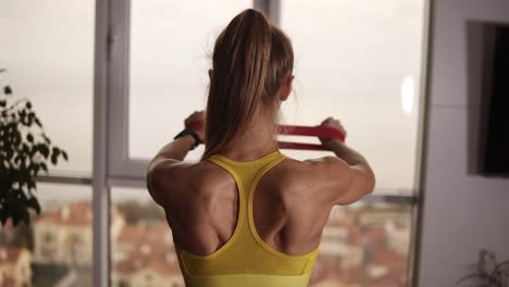 Footage-from-behind-of-a-fit-athletic-woman-doing-pull-ups-using-rubber-band-for-help.-Woman-training-her-back.-Pumping-muscles,-wearing-yellow-sport-top.-Domestic-workouts