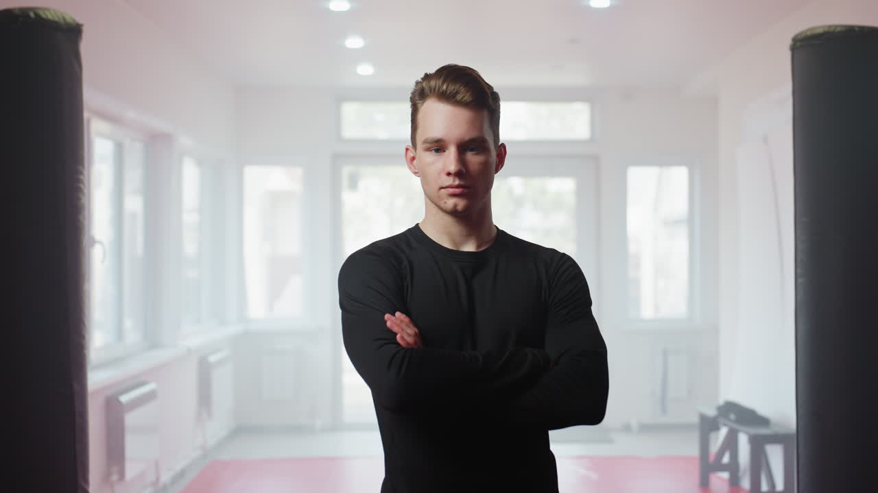Athlete in black sportswear stands focused in gym with punching bags and bright natural light, embodying discipline, strength, and determination while preparing for training session with confident posture