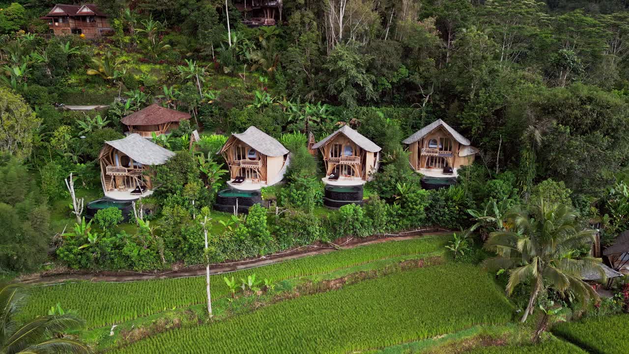 Drone footage of Sidemen Bali featuring bamboo accommodation with infinity pools built above rice fields, tropical vegetation, and rugged hills, offering a clear view of rural island hospitality