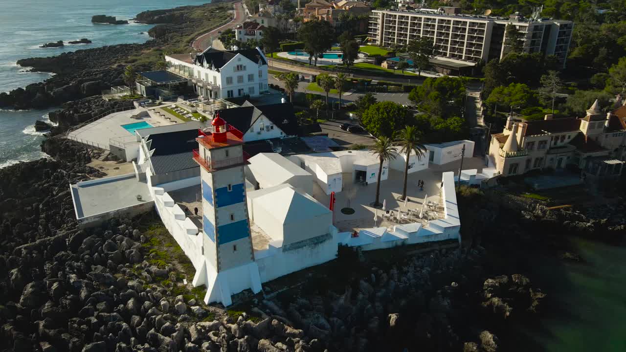 Aerial drone video orbiting around historical Forte de Santa Marta lighthouse and casa de Santa Maria in Cascais Portugal that is on rocky tall reeds where ocean waves hit on shoreline. Sunny summer