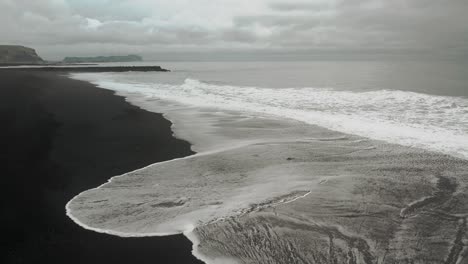 Filmische-Zeitlupe-Aus-Der-Luft,-Meereswellen-Am-Schwarzen-Sandstrand,-Stimmungsvolle-Dunkle-Landschaft,-Vik,-Island