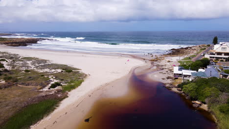 Aerial-toward-beach-over-tannin-rich-waters-of-Onrus-lagoon,-Overstrand